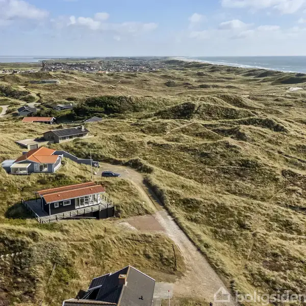 Et hus med orange tag ligger på en bakke omgivet af sandklitter. Der er flere huse i området med udsigt over havet og en klar blå himmel.