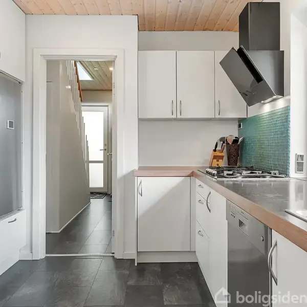 Et moderne køkken med hvide skabe og sorte gulvfliser. En indbygget ovn og gasblus, mens et vindue viser en udendørs have. En dør fører til en anden del af huset.