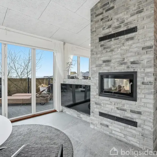 Pejs med glasfront er tændt i moderne stue. Rummet har store vinduer mod terrasse med sofa og liggestol udenfor. Et rundt bord med stol står nær pejsen.