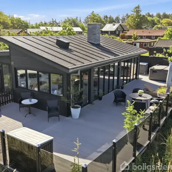 Et sort sommerhus med store vinduer ligger på en rummelig terrasse med moderne havemøbler, omgivet af frodig vegetation og andre huse i baggrunden.