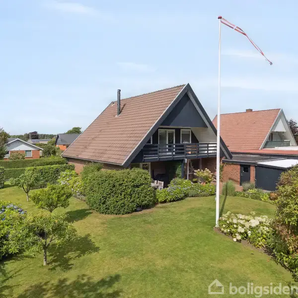 Et A-formet hus med altan står midt i en velplejet have. Huset er omgivet af grønne buske og træer. En flagstang med Dannebrog vajer i forhaven.