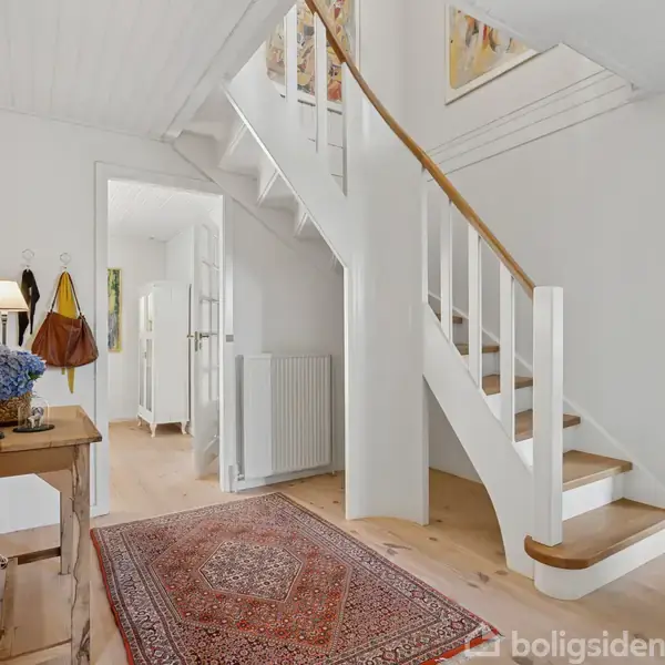 En trætrappe med hvidt gelænder står i et lyst rum med lyse trægulve. Bord til venstre med blomster og lamper, tæppe på gulvet, døråbning til andre rum.