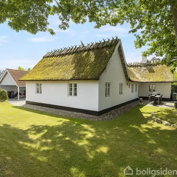 Et hvidt bindingsværkshus med stråtag står på en velplejet græsplæne omkranset af træer og buske. En terrasse med havemøbler er placeret til højre for bygningen.