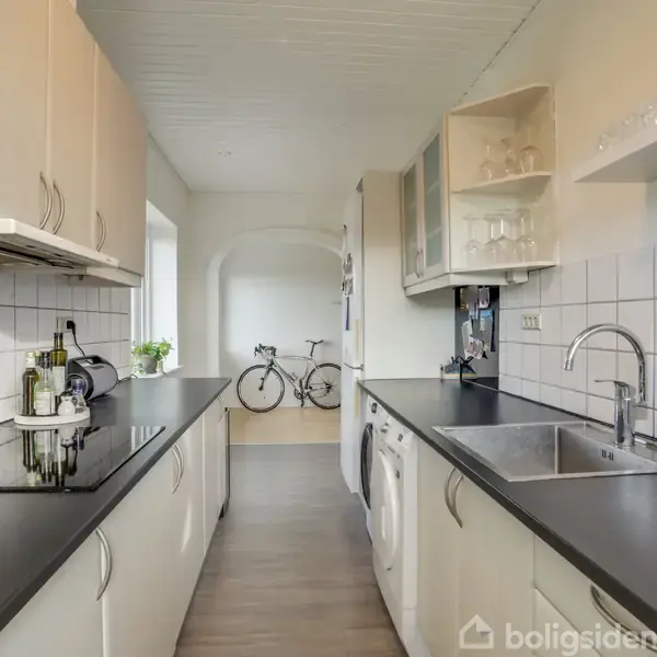 Et moderne køkken med sorte bordplader strækker sig langs væggene. Vasken og komfuret er indbygget. En cykel står i et tilstødende rum bag en bueåbning.
