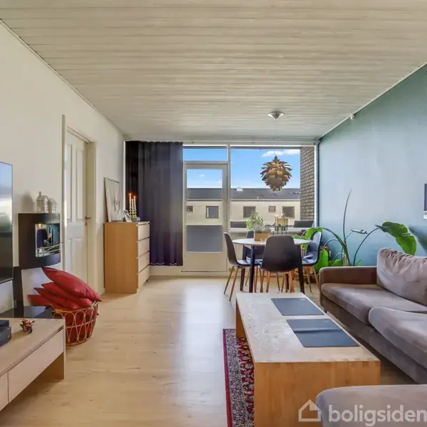 En stue med en sofa til højre, et fladskærms-tv til venstre, og et spisebord nær bagvinduet. Grøn plante og sofabord tilføjer detaljer til rummet.