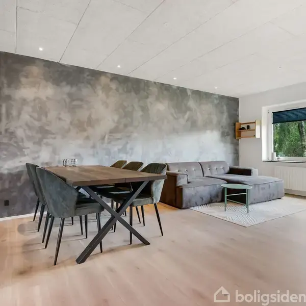 Moderne spisebord med seks stole står foran en grå væg. Ved siden af er en grå hjørnesofa i en lys stue med trægulv og vinduer med udsigt til træer.