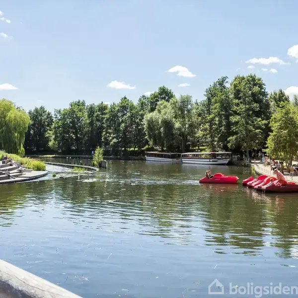 En stille sø med to røde paddle-både, omkranset af træer og en promenade med caféer, under en klar blå himmel.