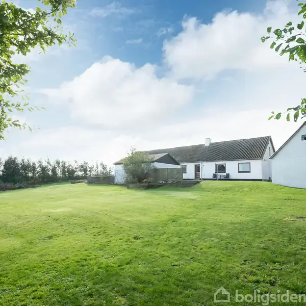 Hus ligger fredfyldt på en stor, lys grønt græsplæne omgivet af træer og buske under en blå himmel med skyer.