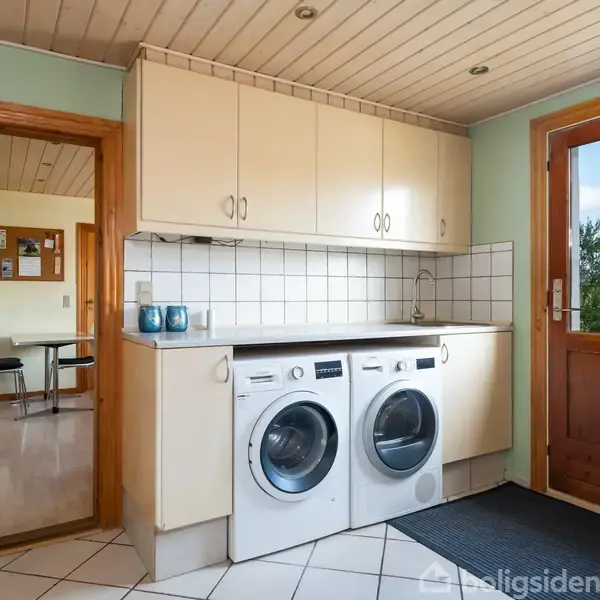 Vaskemaskine og tørretumbler står side om side under en hvid bordplade i et lysegrønt bryggers med hvide overskabe. En dør til højre fører ud til en have.