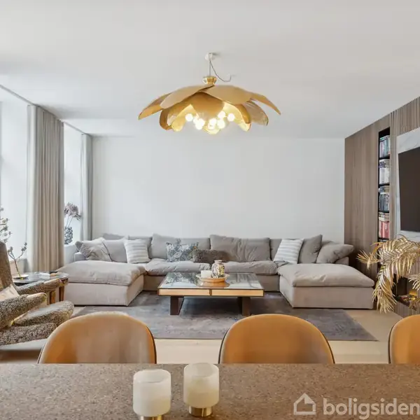 Grå sofa står i moderne stue, flankeret af store vinduer og planter. Rundt sofabord har dekorationer. Lamper hænger fra loftet. Væghængt TV ses mod bagvæg.