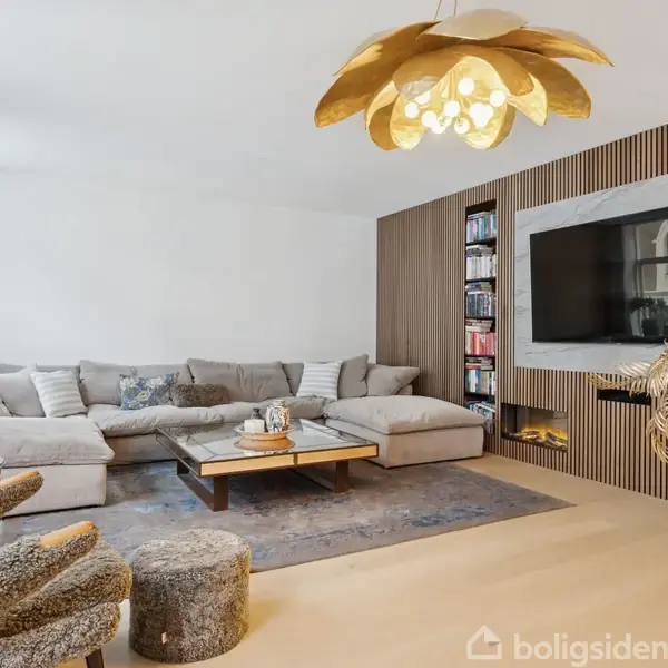 En stor, grå sofa står i et moderne opholdsområde med en fladskærm på væggen, en unik gul lampe i form af et blad og dekoreret med planter og et tæppe.