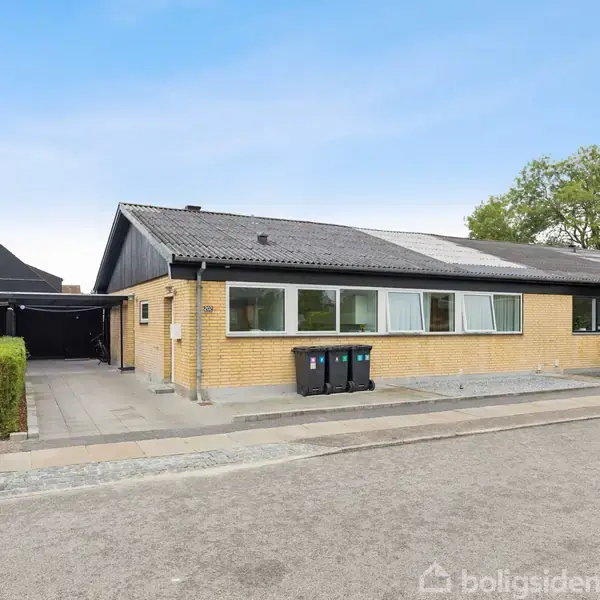Et gulstenshus med fladt tag står stille på en stille vej. Tre skraldespande er linet op langs husmuren. På højre side er der en indkørsel og en carport.