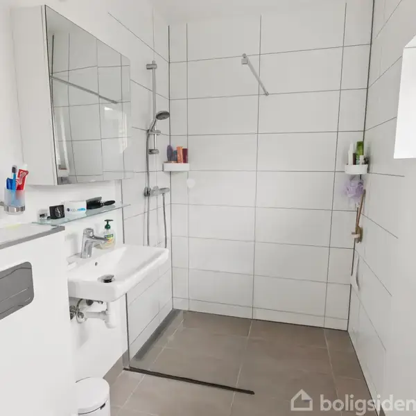 Et moderne badeværelse med brusekabine til venstre, hvidt flisegulv og vægge. Der er en håndvask med spejl og toilethylde med toiletartikler. En lille vindue findes til højre.