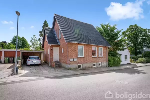 Rødt murstenshus med sort tag står langs en stille villavej. En hvid bil er parkeret i en overdækket carport til venstre. Omgivet af træer og grønne buske.