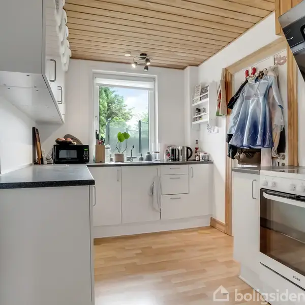 Køkken med lyse træskabe og en ovn til højre, mikroovn til venstre, vindue i midten. Tilbehør som kedel og opvaskemiddel. Lyse trægulve og trælofter skaber en hyggelig atmosfære.