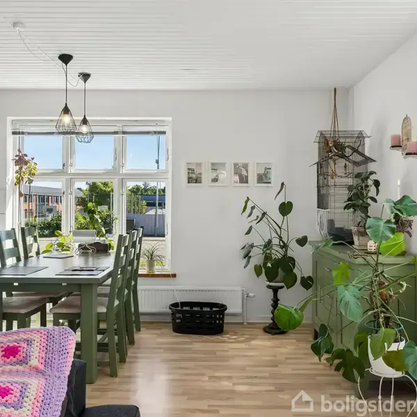 En lys spisestue med et grønt spisebord og seks stole. Planter dekorerer rummet, inklusive store ved vinduet og skænken. Et tæppe med mønstre ligger på sofaen.
