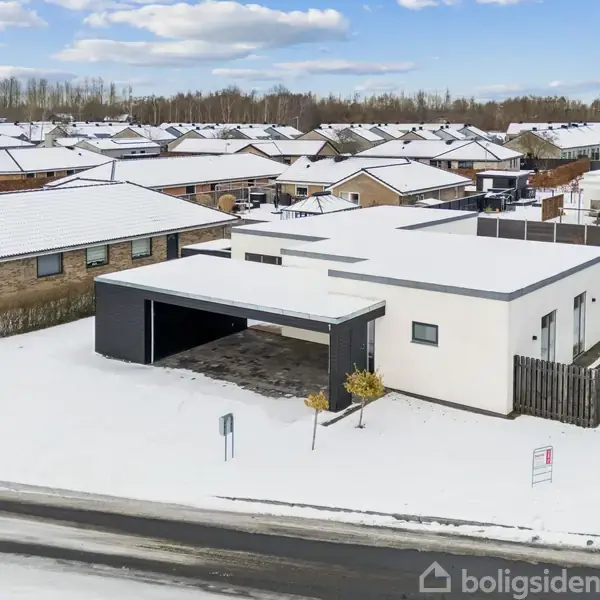 En moderne, hvid bygning med fladt tag står i et sneklædt boligområde. Omkringliggende huse har skrå tage, og en sort indkørsel fører til carporten ved siden af huset.