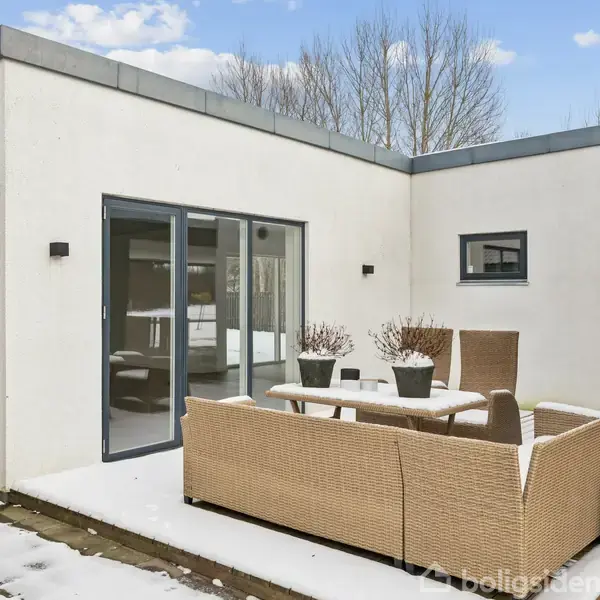 Terrassemøbler af fletmateriale står udenfor et moderne, hvidt hus med store vinduer. Sne dækker jorden, og himlen er let overskyet. Planter i krukker står på bordet.