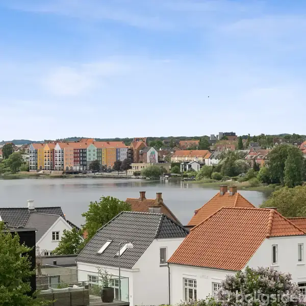 Murstens- og teglhuse ligger fredeligt i forgrunden med udsigt til en sø. I baggrunden ses flerfarvede lejlighedsbygninger og et grønt landskab under en blå himmel.