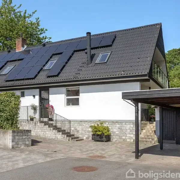 Huset er hvidt med en sorttagterrasse, solpaneler installeret. Trappe leder til hoveddøren. Carport på højre side med affaldsbeholdere. Grønne omgivelser bagved og ved siden af.