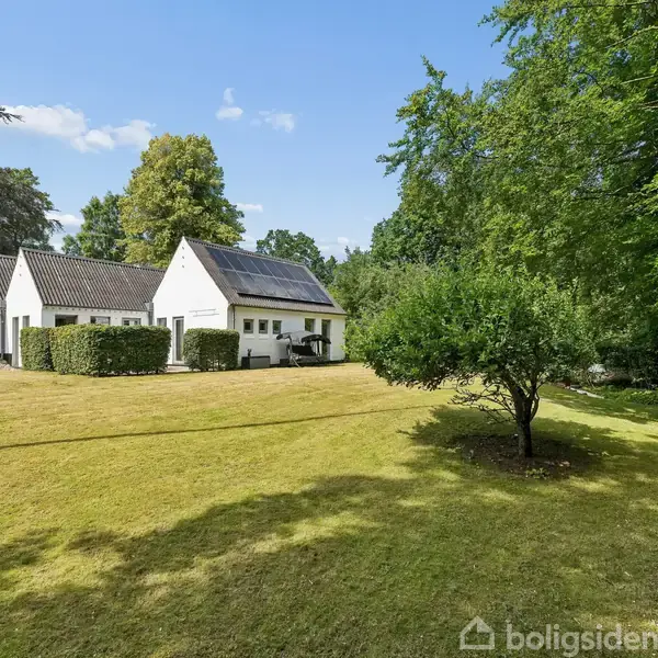 En hvid villa med solpaneler på taget står i en stor have omringet af grønne træer og buske under en blå himmel.