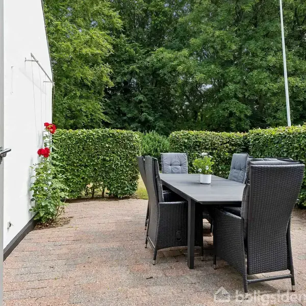 Terrassebord med seks sorte stole står på en flisebelagt terrasse. I baggrunden ses hække og buske, der omgiver området, samt en hvid bygning til venstre.