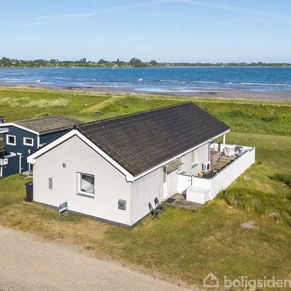 Et hvidt hus med mørkt tag står ved vejen, omgivet af græsplæne. Det ligger nær en strand med udsigt over havet under en klar blå himmel.