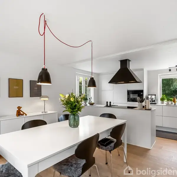Et moderne køkken med hvidt interiør. Bord med sorte stole, vase med blomster, hængelamper og udsigt til grønne træer udenfor. Hyggelig indretning med vægdekorationer.