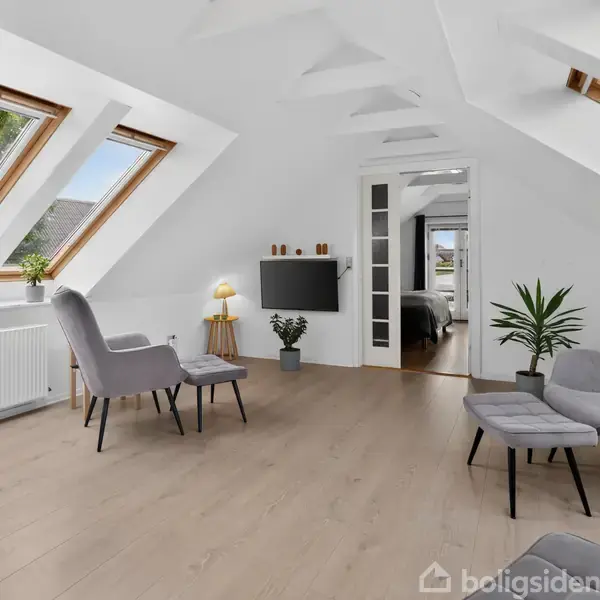 En lys stue med skrålofter har grå stole og et lille bord. Sollys strømmer ind gennem de store ovenlysvinduer. En fladskærm hænger på væggen med en plante ved siden af.