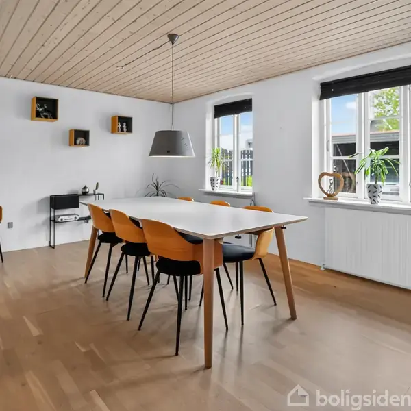 Et spisebord med seks stole står i et lyst, moderne rum. Store vinduer lader dagslys ind, og en grå lampe hænger over bordet. Planter står ved vinduet.