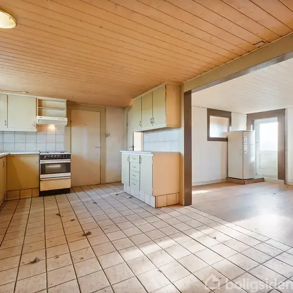 Køkken med gule skabe står tomt. En komfur og emhætte ses ved væggen. Rummet er stort med flisegulv og åbner mod en tilstødende stue med trægulv.