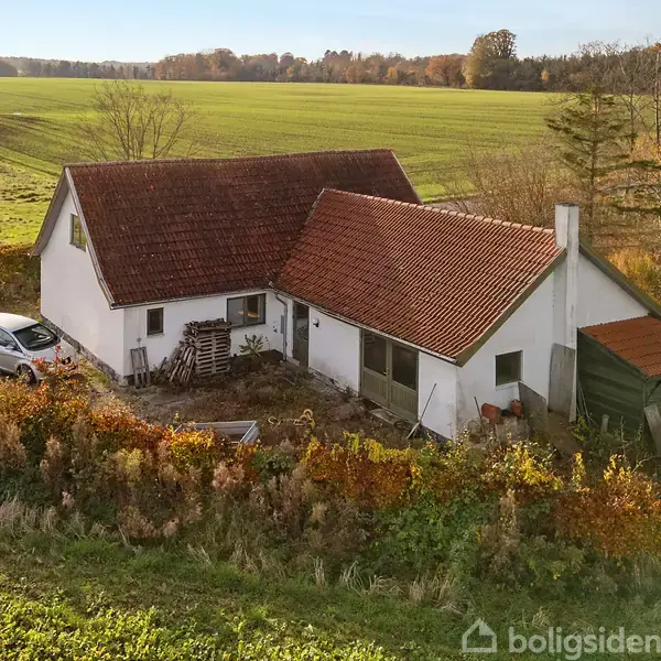 Hus med rødt tegltag ligger i landligt område. En bil holder parkeret ved siden af huset, omgivet af grønt græs og skove i baggrunden.
