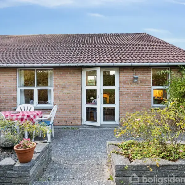 Murstenshus med rødt tegltag, har en terrasse foran med bord og stole. Omgivet af grønne planter og små blomsterbede. Skyer og blå himmel ses i baggrunden.