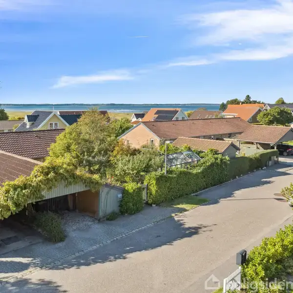 Huse med røde tegltage står stille langs en stille, solbeskinnet vej. I baggrunden er der udsigt til havet og en klar blå himmel.