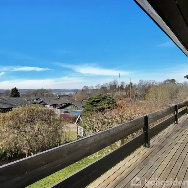 En lang træterrasse strækker sig langs et hus med udsigt over frodige haver og hustage, med en blå himmel i baggrunden. En hvid spisebordssæt står på terrassen.