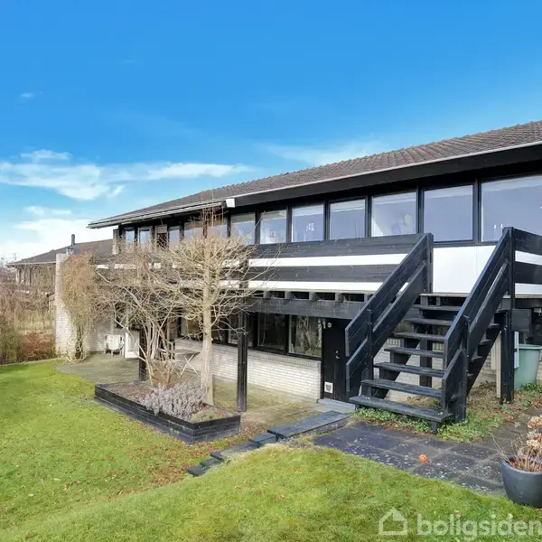 Et moderne hus med store vinduer og en udendørs trappe forbinder to etager. Det ligger i en velholdt have med græs og planter, under en klar blå himmel.