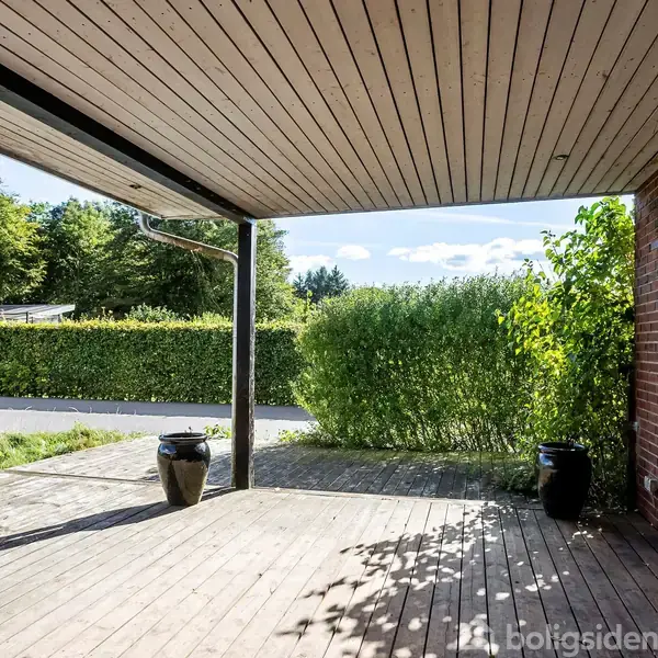 En overdækket terrasse med træbrædder og to sorte krukker. Omgivet af grønne buske og en græsplæne foran en hæk, perfekt til udendørs afslapning.