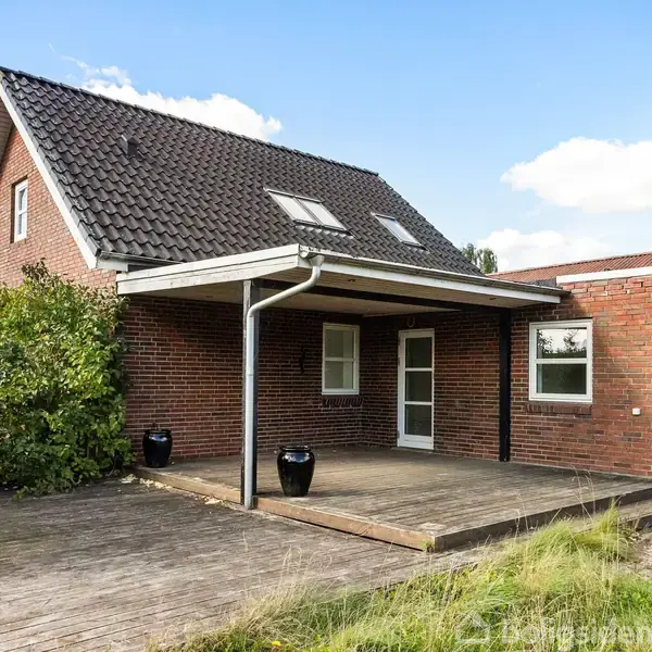 Et rødt murstenshus med sort tegltag står stille i en have med vildtvoksende græs. En stor træterrasse med overdækning er foran huset, mens buskads omkranser siden.