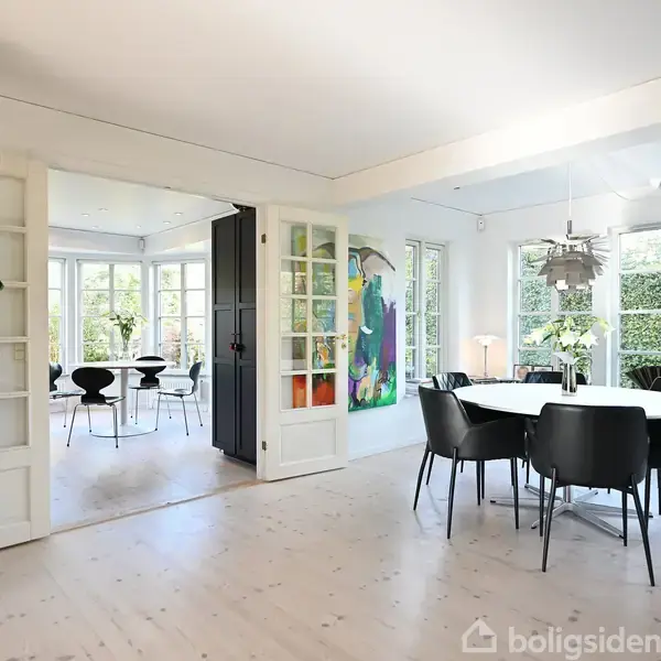 Et rundt spisebord med sorte stole står i en lys stue. Store vinduer og en plante i hjørnet bidrager til en rummelig, moderne atmosfære.