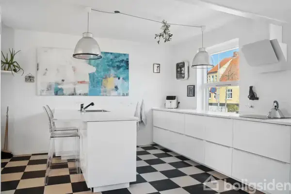 Et lyst køkken med et sort-hvidt skakternet gulv. To hængelamper over en køkkenø med klare barstole. Et vindue viser husfacade. Abstrakt kunst hænger på væggen.