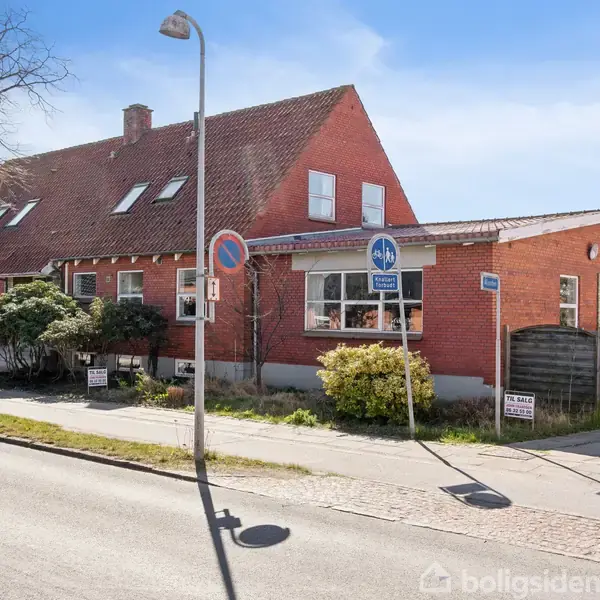 Et rødt murstenshus med en skråtag udfører boligtjeneste i en byzone, omgivet af fortov og vejskilte. Tekst: "P-Plads Kun For Ejendommens Kunder."