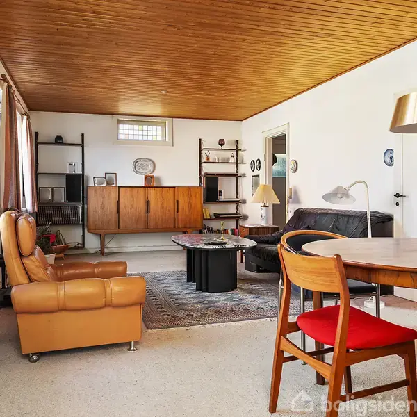 Stue indrettet med læderlænestol, sofabord og sort sofa, placeret på tæppe. Rundt spisebord med røde stole står nær vindue med gardiner. Træloft og hvide vægge skaber en hyggelig atmosfære.