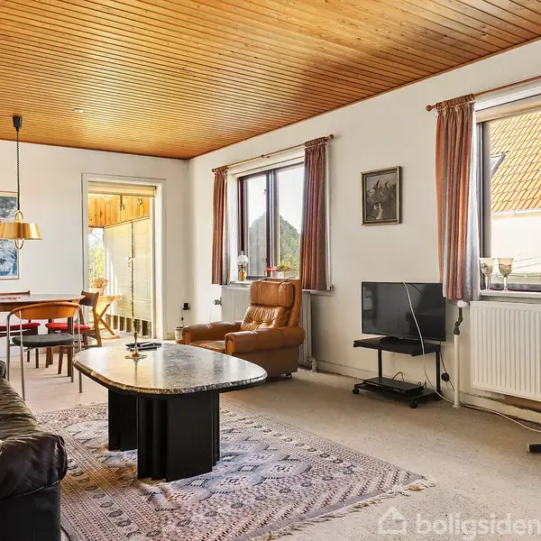 En stue med en sort sofa, et ovalt sofabord og en lænestol. Spisebord står i det lyse hjørne ved et vindue. Trægulv og loft skaber en varm atmosfære. Ingen tekst.
