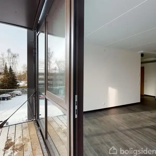 Balkon med glasdør, åben til indendørs stue med mørkt trægulv; udenfor ses sneklædt landskab med træer og bygninger i baggrunden.