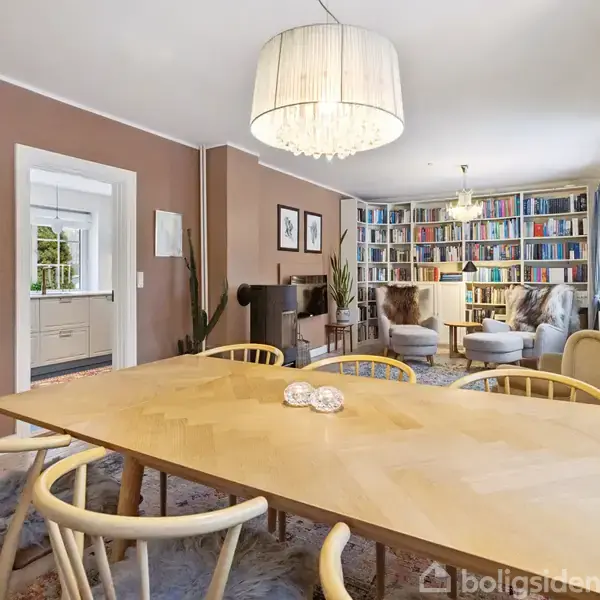 Langt spisebord med lyse træstole står i en elegant stue. Væggene er brune, bøger fylder reolerne i baggrunden, og et lyst tæppe dækker gulvet under en stor lysekrone.