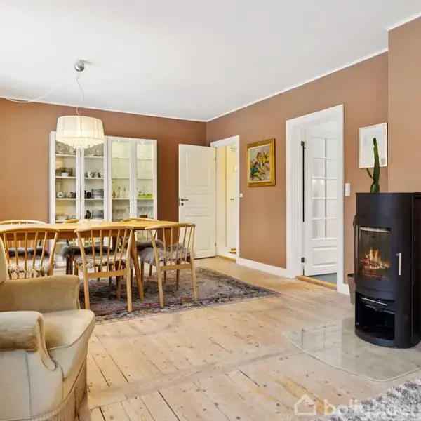 En brændeovn står tændt i stuen, omgivet af en beige lænestol, et spisebord med stole, et vitrineskab, og vægge i brunlige nuancer. En kurv og tæppe er på gulvet.