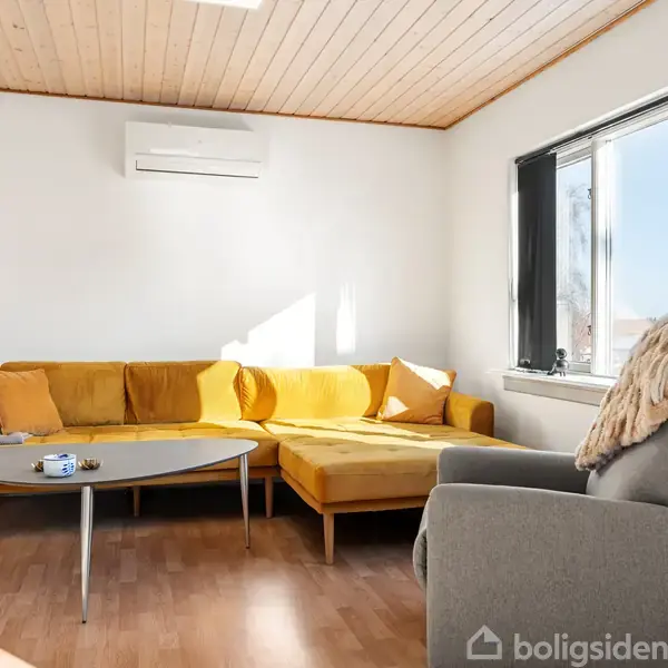 En sennepsgul sofa står ved et stuebord med en kaffekop i en lys stue. Ved siden af er en grå lænestol nær et vindue med udsigt til blå himmel.