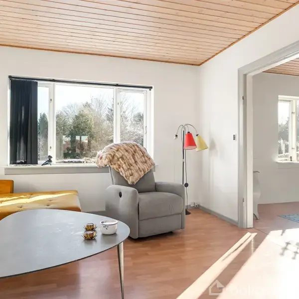 En stue med en gul sofa ved siden af et gråt lænestol med tæppe, står foran et stort vindue. Et trekantet sofabord står foran sofaen.