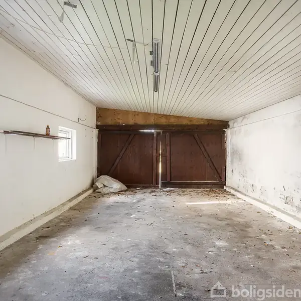Garage med tomt interiør, hvor mørke træporte dominerer i enden. Der er støvede betongulve, en hvidmalet træloft, samt to små vinduer på den ene væg.