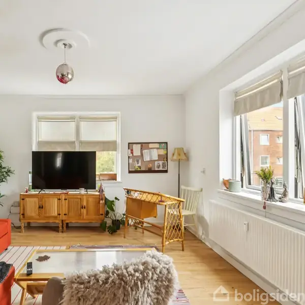 En stue med en rød sofa står ved vinduet. Et træbord med en TV-enhed udfylder rummet. Der er også en stol og plante ved siden af. Spejl og opslagstavle pryder væggene.